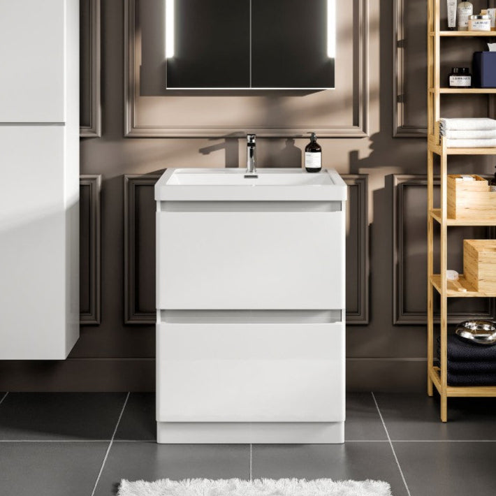 Modern white single sink bath vanity with acrylic top and integrated sink, featuring a two-drawer design, placed in a bathroom setting with dark walls, a mirrored medicine cabinet, and a wooden shelf with toiletries.
