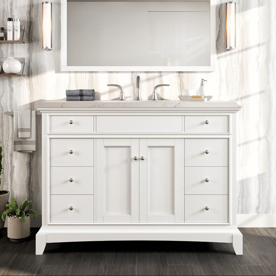 A white single sink bath vanity with a Carrara quartz top, mirrored by a rectangular mirror, with lighting fixtures on either side of the mirror. The vanity has multiple drawers and a central cabinet, with a porcelain sink installed on top.
