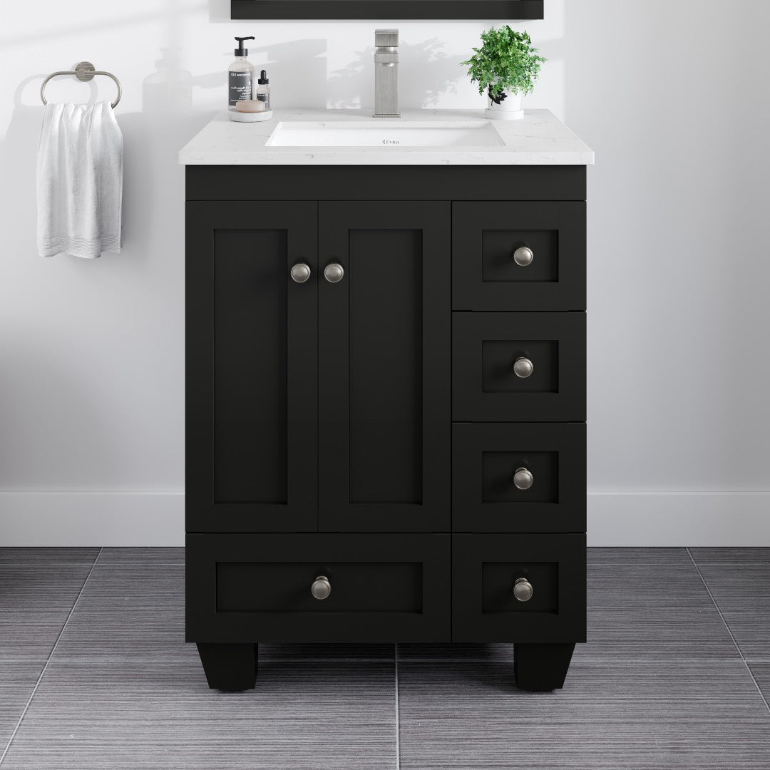 A black, single sink bathroom vanity with a white Carrara Quartz countertop and silver brushed nickel hardware. The vanity has multiple drawers and a lower cabinet, set against a white tiled wall with a plant to the side.
