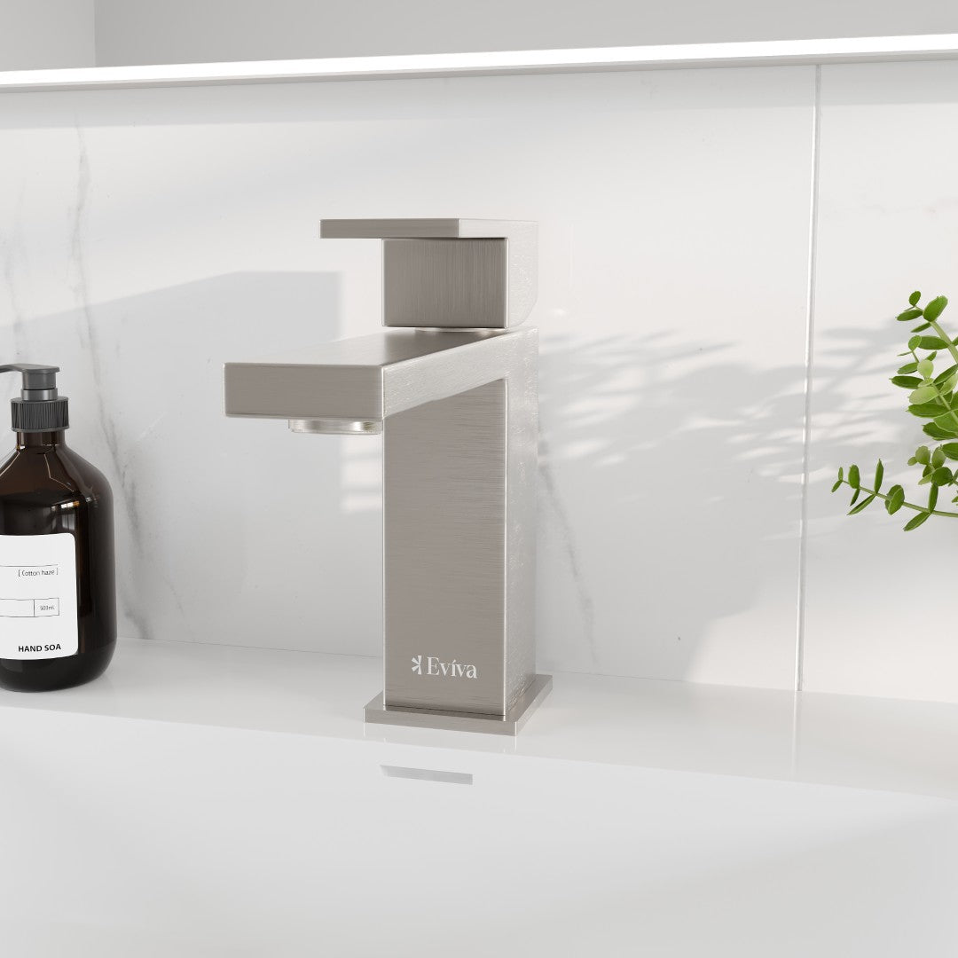 Brushed nickel single-handle waterfall bathroom faucet with a deckplate, displayed in a kitchen setting with a bottle of soap and a plant in the background.