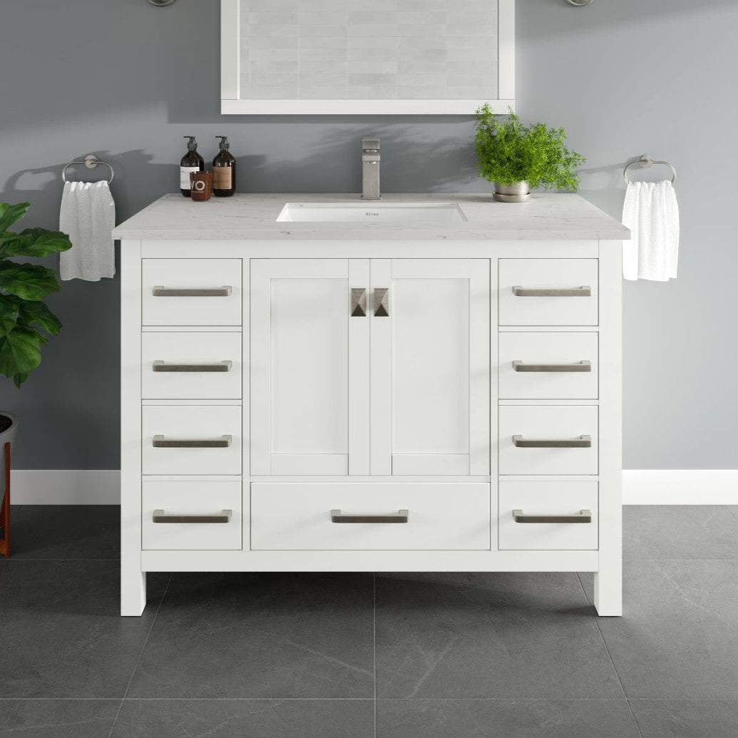 A white single sink bath vanity with a Carrara quartz top and undermount porcelain sink, featuring silver and gold hardware, placed in a bathroom setting with decorative items and a plant in the background.