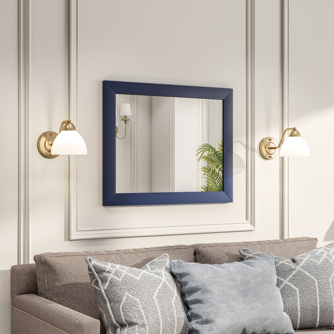 A square, dark blue framed mirror hangs on a light-colored wall above a couch with patterned pillows showing two sconce lights on both side