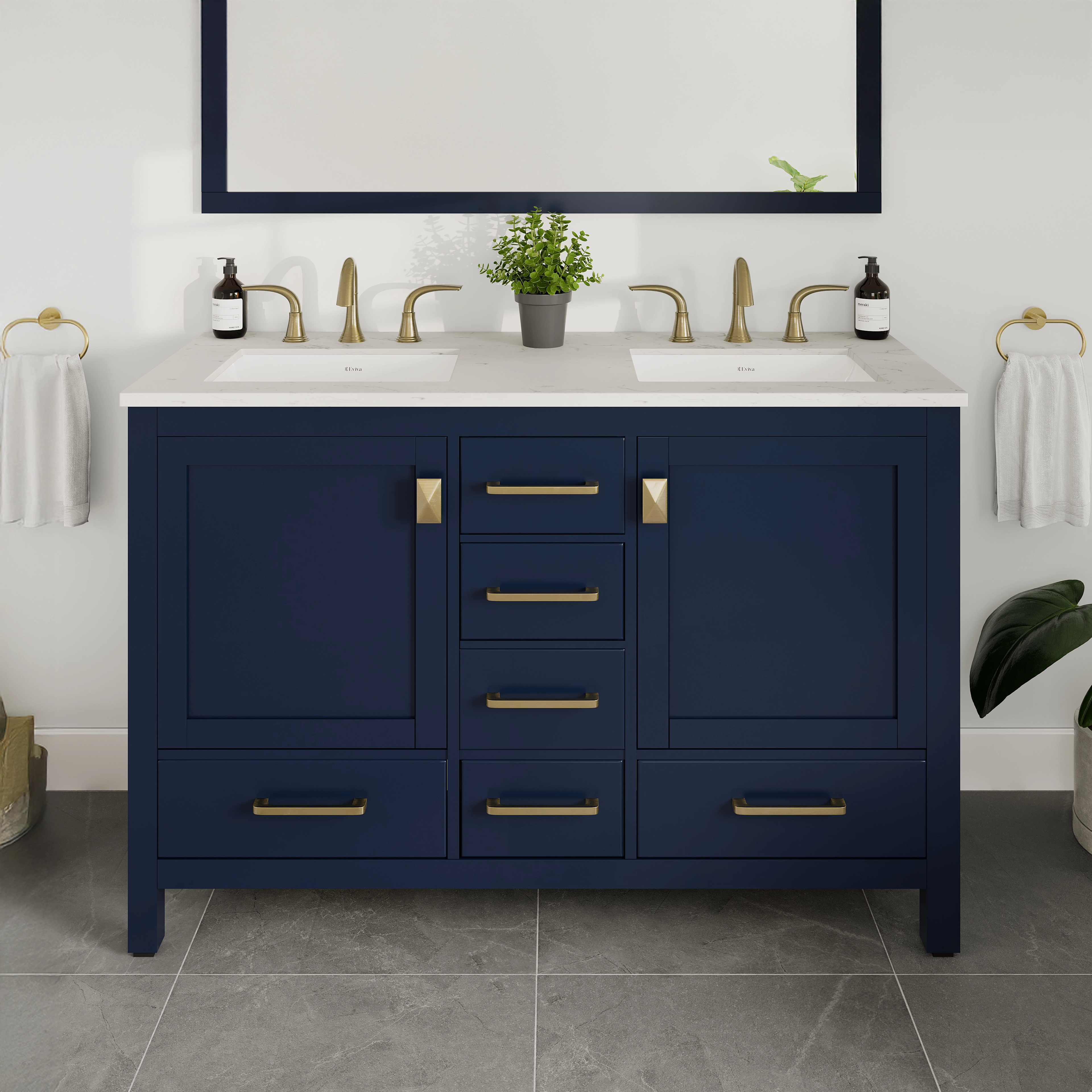 A blue double sink bath vanity with gold and silver hardware, a white Carrara quartz top, and undermount porcelain sinks. The vanity is positioned against a white wall on a grey tiled floor, with a mirror above the sink and decorative plants to the sides.