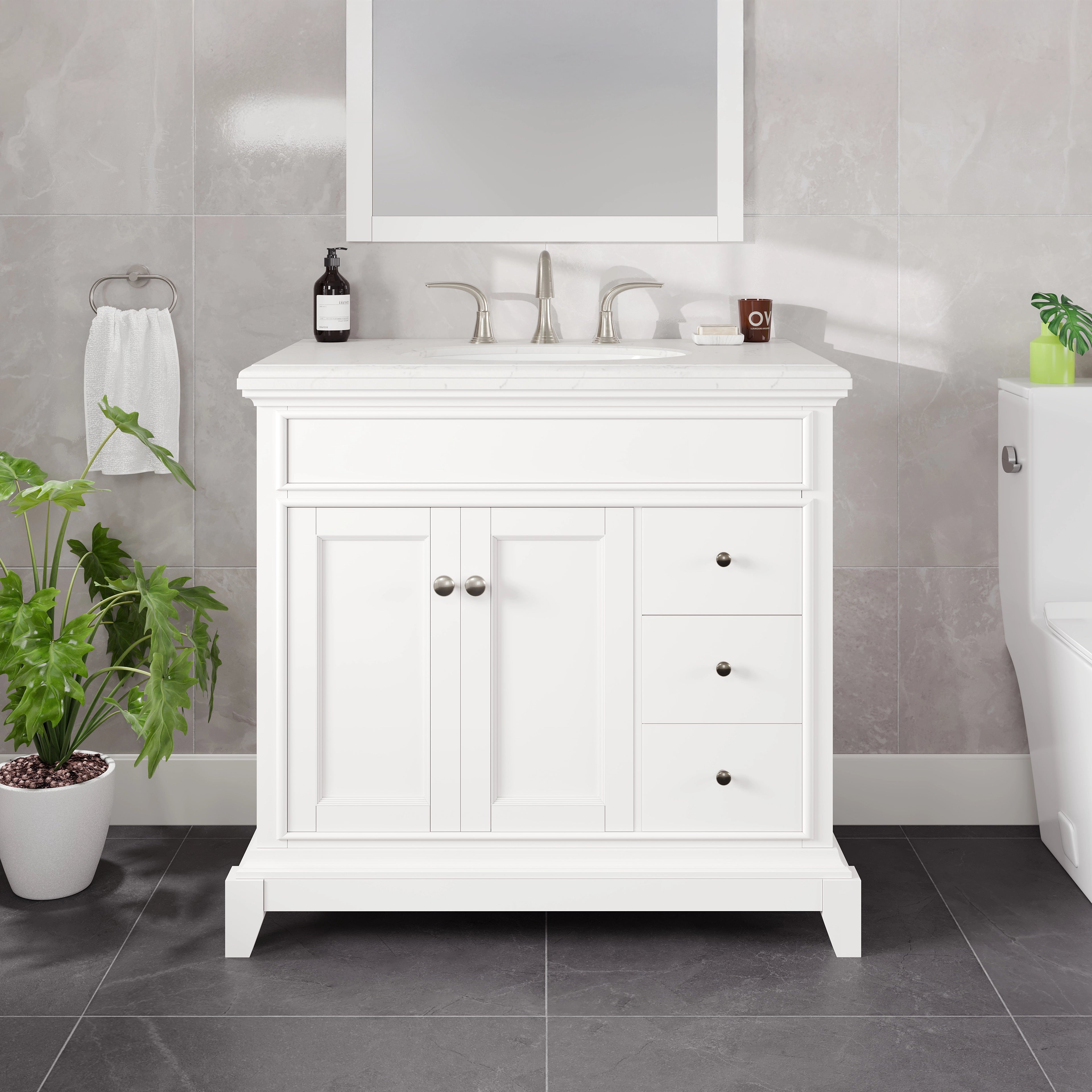 A white single sink bathroom vanity with a Carrara Quartz countertop, undermount porcelain sink, and two doors. The vanity is displayed in a bathroom setting with a mirror above the sink, a plant to the side, and a towel hanging on the wall.