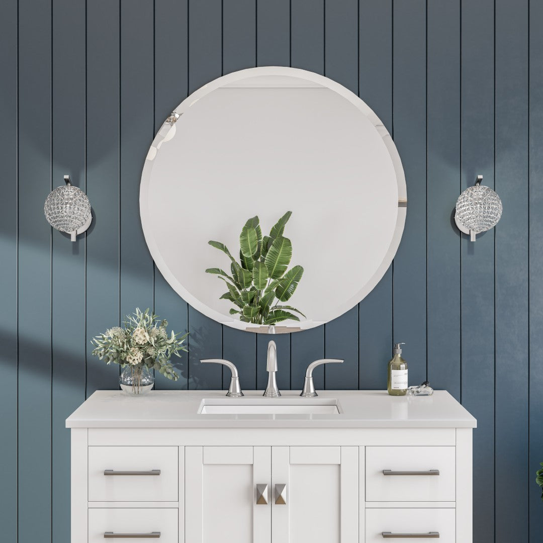 A round, frameless mirror with a silver aluminum frame mounted on a blue wall above a white vanity with a glass mirror surface reflecting a plant and light fixture.