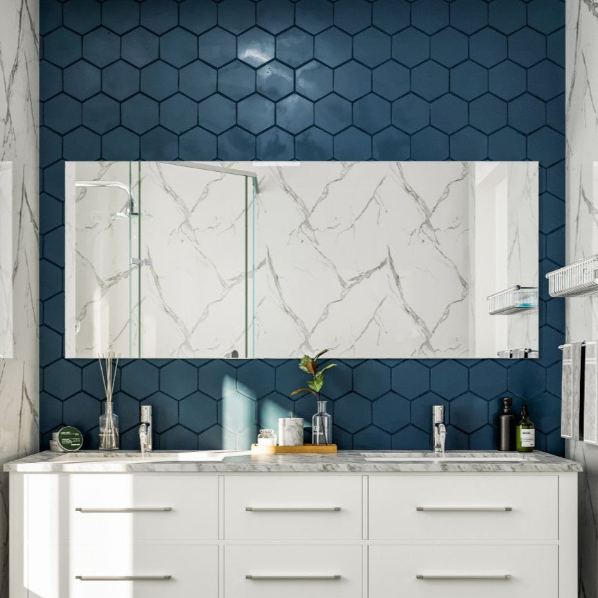 A rectangular, frameless mirror with beveled edges mounted on a wall with white marble tiles, above a white vanity with marble countertop.