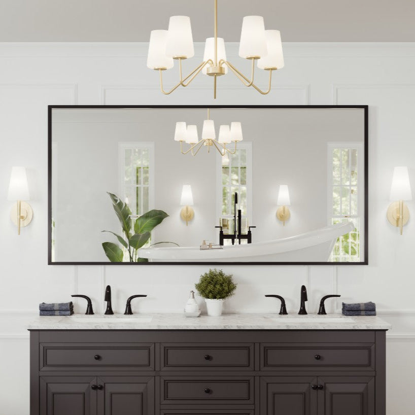 A rectangular black metal framed mirror mounted above a bathroom vanity with dual sinks, surrounded by decorative elements such as plants and a chandelier.