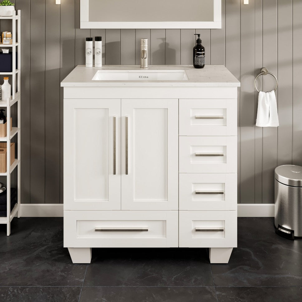 A white single sink bath vanity with a Carrara quartz top and undermount porcelain sink, featuring a white finish and stainless steel hardware, placed in a bathroom setting with dark flooring and wall tiles.