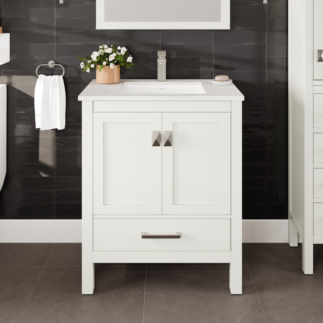 A white single sink bath vanity with a Carrara quartz top and undermount porcelain sink, displayed in a bathroom setting with black tiled wall and floor.