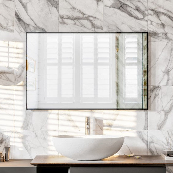 A rectangular black metal framed mirror mounted on a wall with marble tiles, above a modern bathroom vanity with a white sink.