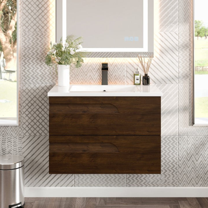 Modern wall-mounted single sink bath vanity with rosewood finish, white porcelain top, and integrated sink, illuminated by a mirror with built-in lights. The image also includes a decorative plant, a mirror reflection of a wooden door, and a trash bin.