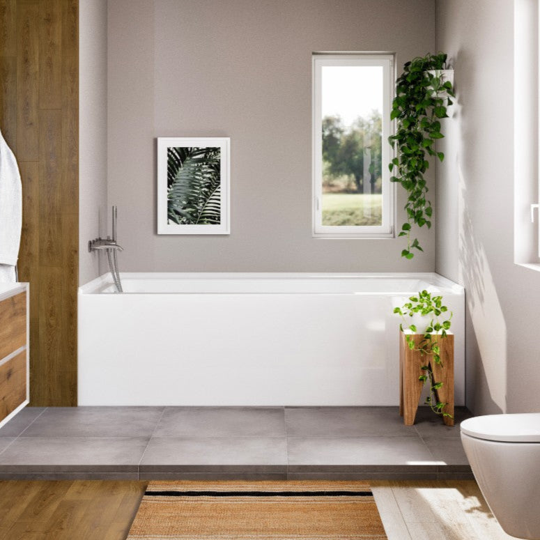 A glossy white acrylic freestanding bathtub installed in a bathroom with a chrome overflow drain, positioned against a window, with a plant decoration on the windowsill.