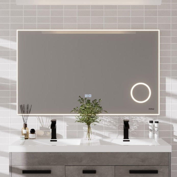 A modern rectangular LED bathroom mirror with a chrome aluminum framed design, mounted on a wall above a vanity. The mirror has a built-in magnifier circle and touch sensor activation features.